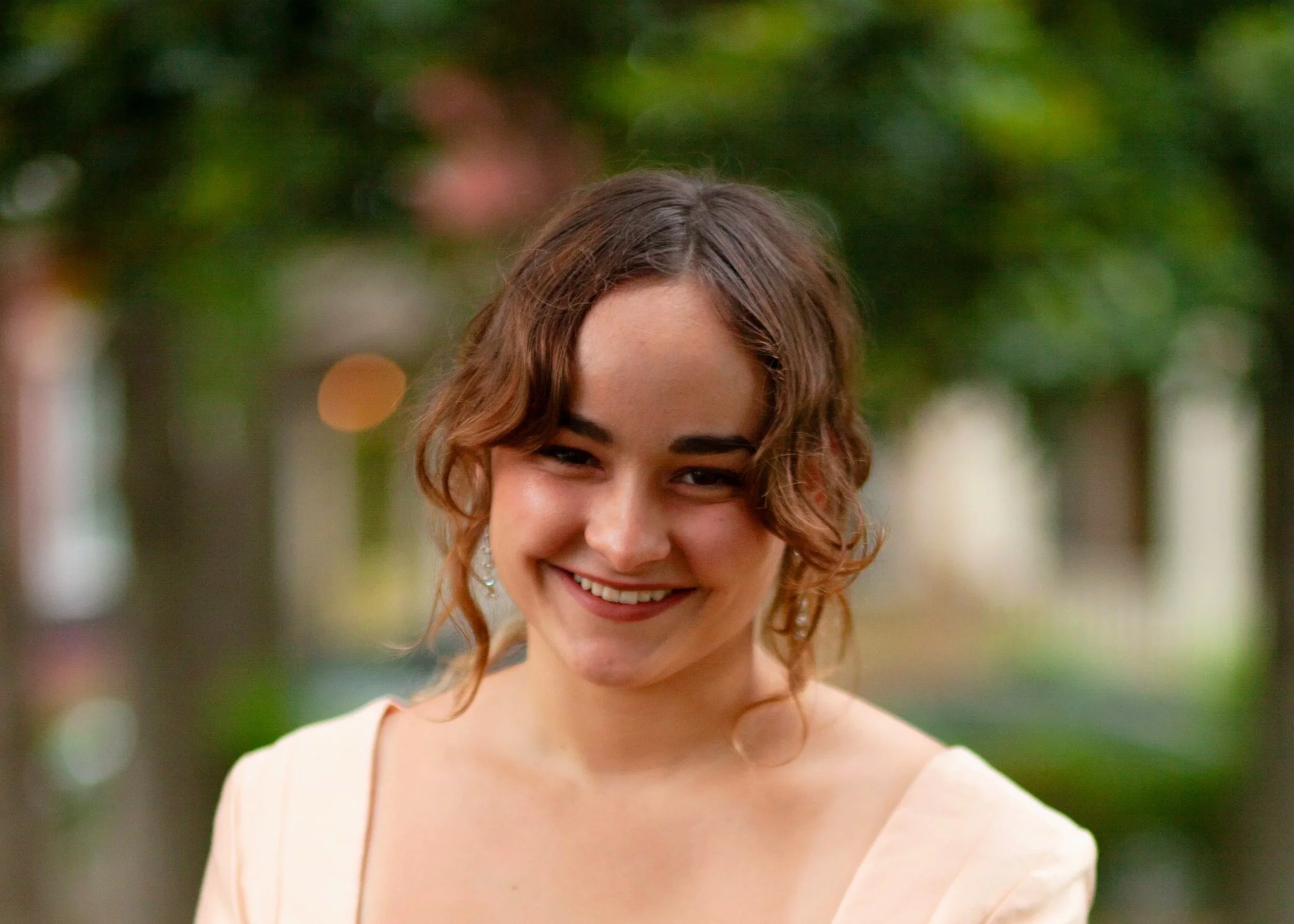 A smiling woman with curly brown hair, wearing a light-colored dress and earrings, standing outdoors with a blurred background of trees and lights.