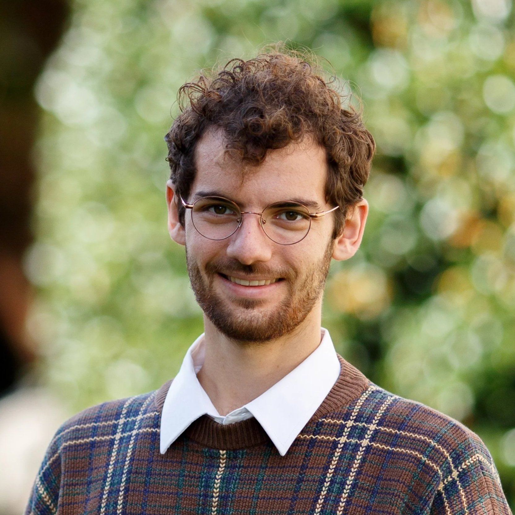A young man with curly brown hair, glasses, and a beard, smiling outdoors with a blurred background of green trees.