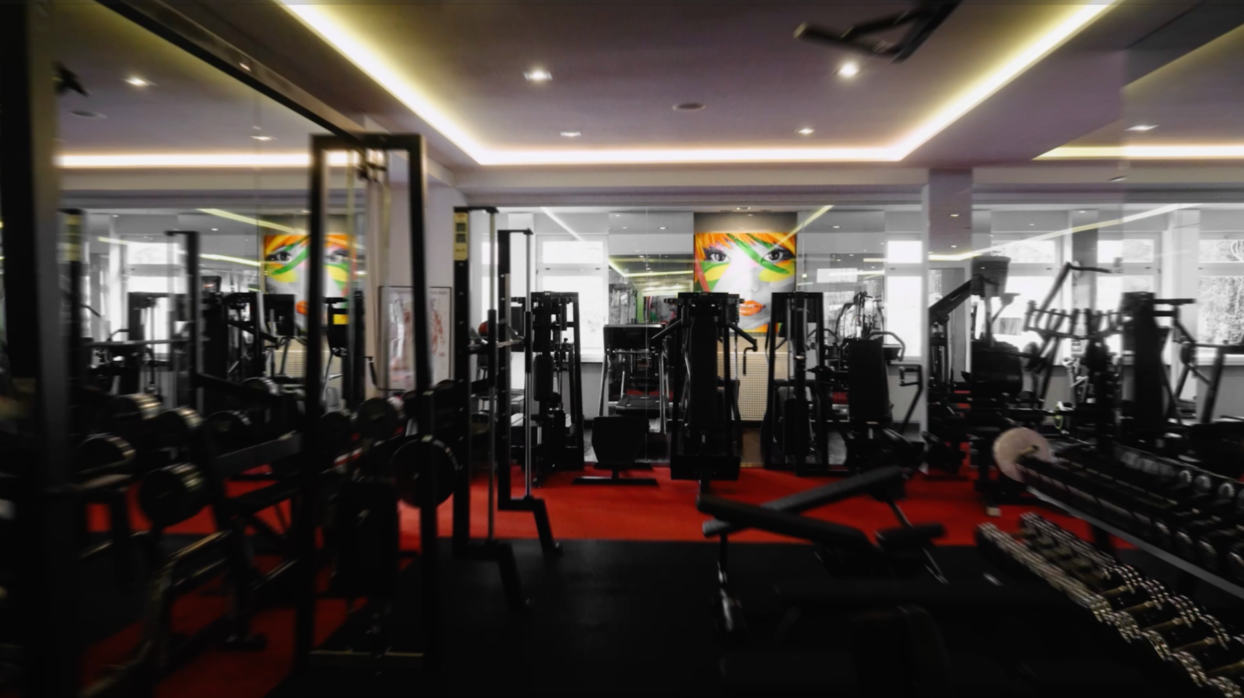 Innenraum eines Fitnessstudios mit verschiedenen Trainingsgeräten, bemaltem Wandbild einer Frau mit farbigen Gesichtszügen und verzierten Augen. Helle Beleuchtung, große Fenster, roter und schwarzer Boden.