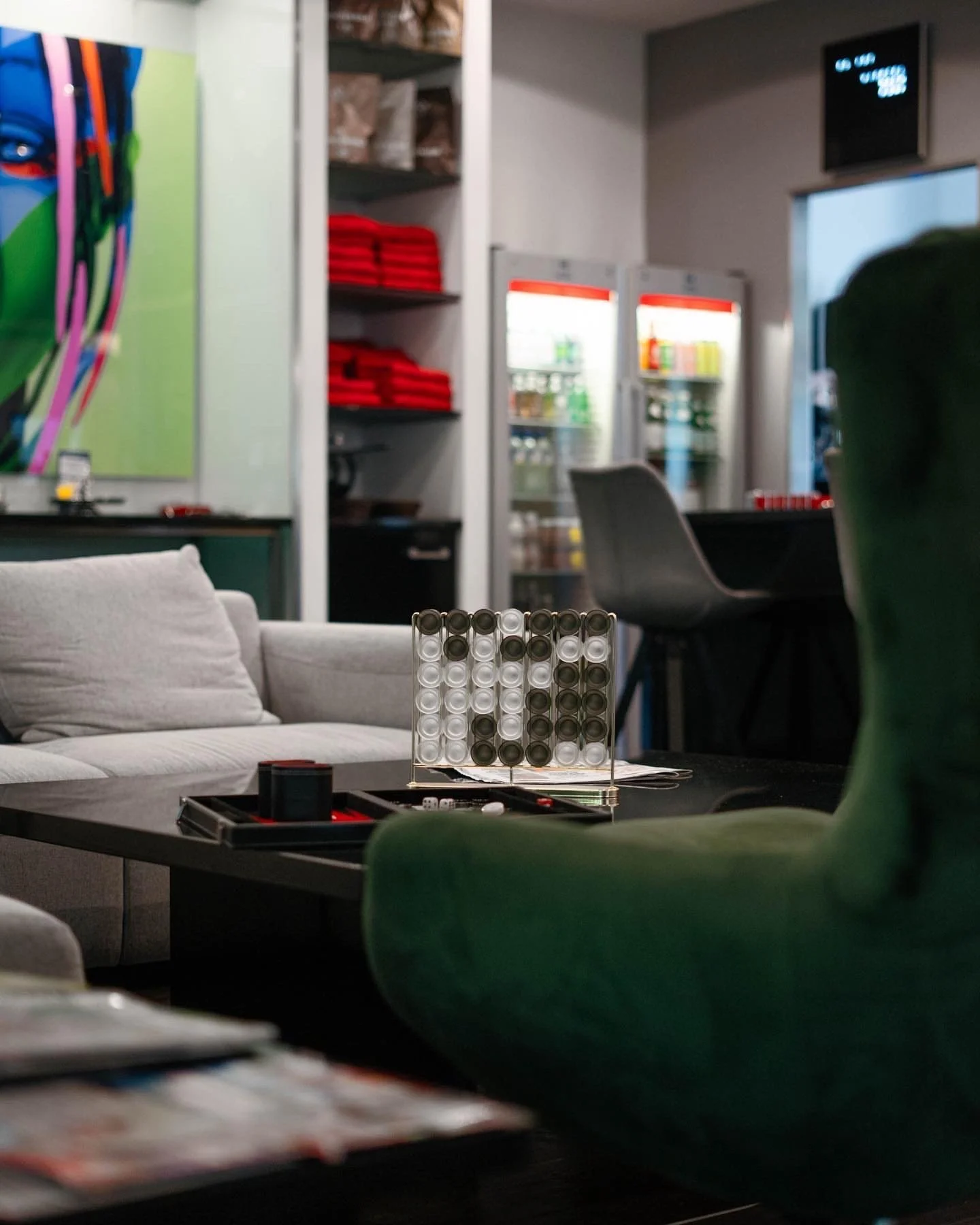 Interior of a lounge or game room with a coffee table, a large sofa, and a green armchair. On the coffee table is a board game with a grid of black and white pieces. In the background are shelves with red towels, a display fridge with beverages, and public seating, including chairs and a couch.