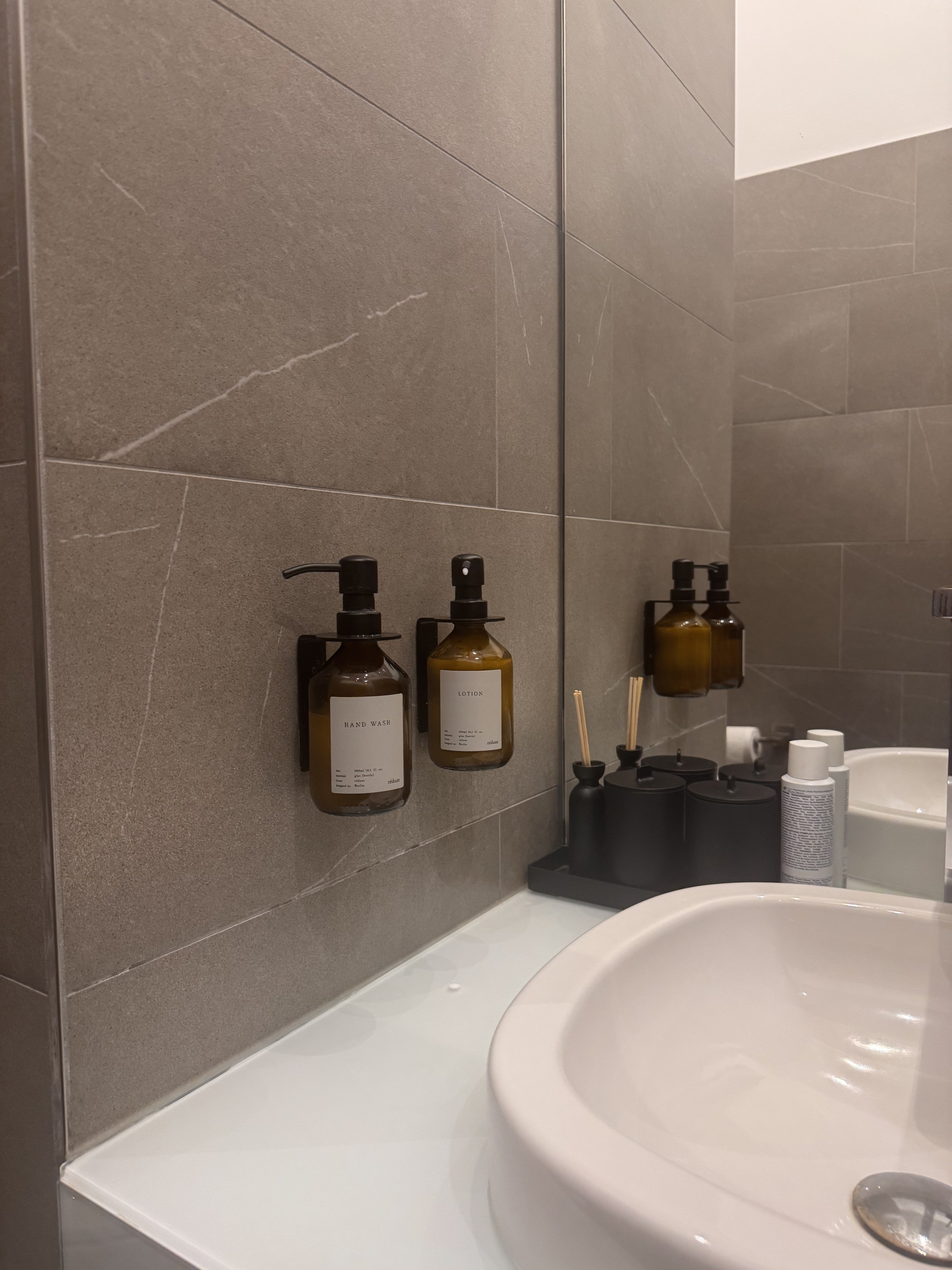 A bathroom sink with a mirror above it, mounted soap and lotion dispensers on the wall, and a black tray with candles and reed diffusers.