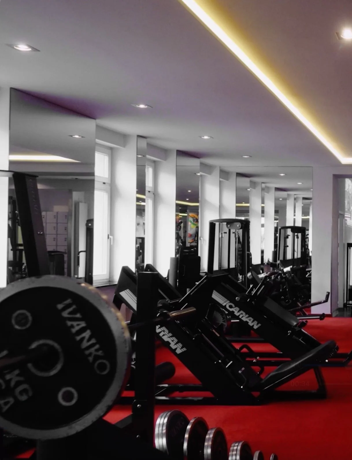 Interior of a modern gym with workout equipment such as weight plates and machines on a red floor, illuminated by ceiling lights and natural light from windows.