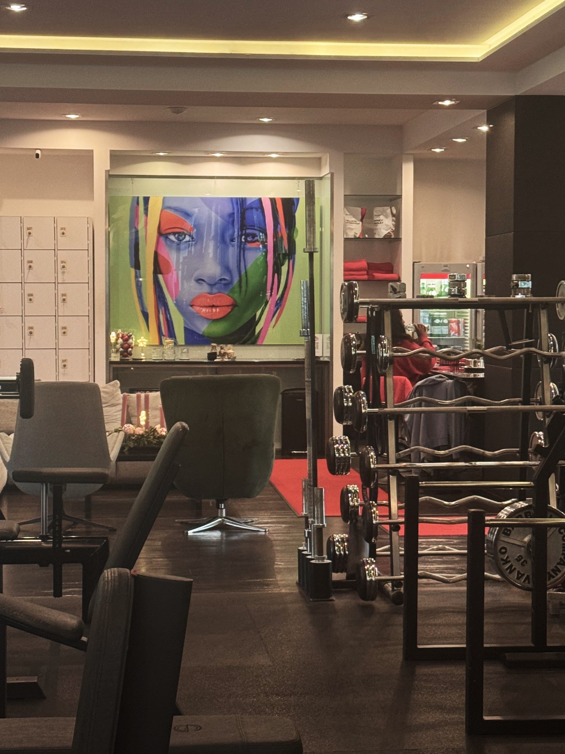A gym with a colorful artwork of a woman's face on the wall, gym equipment including dumbbells and a weight rack, and sitting areas with chairs and a table in the foreground.