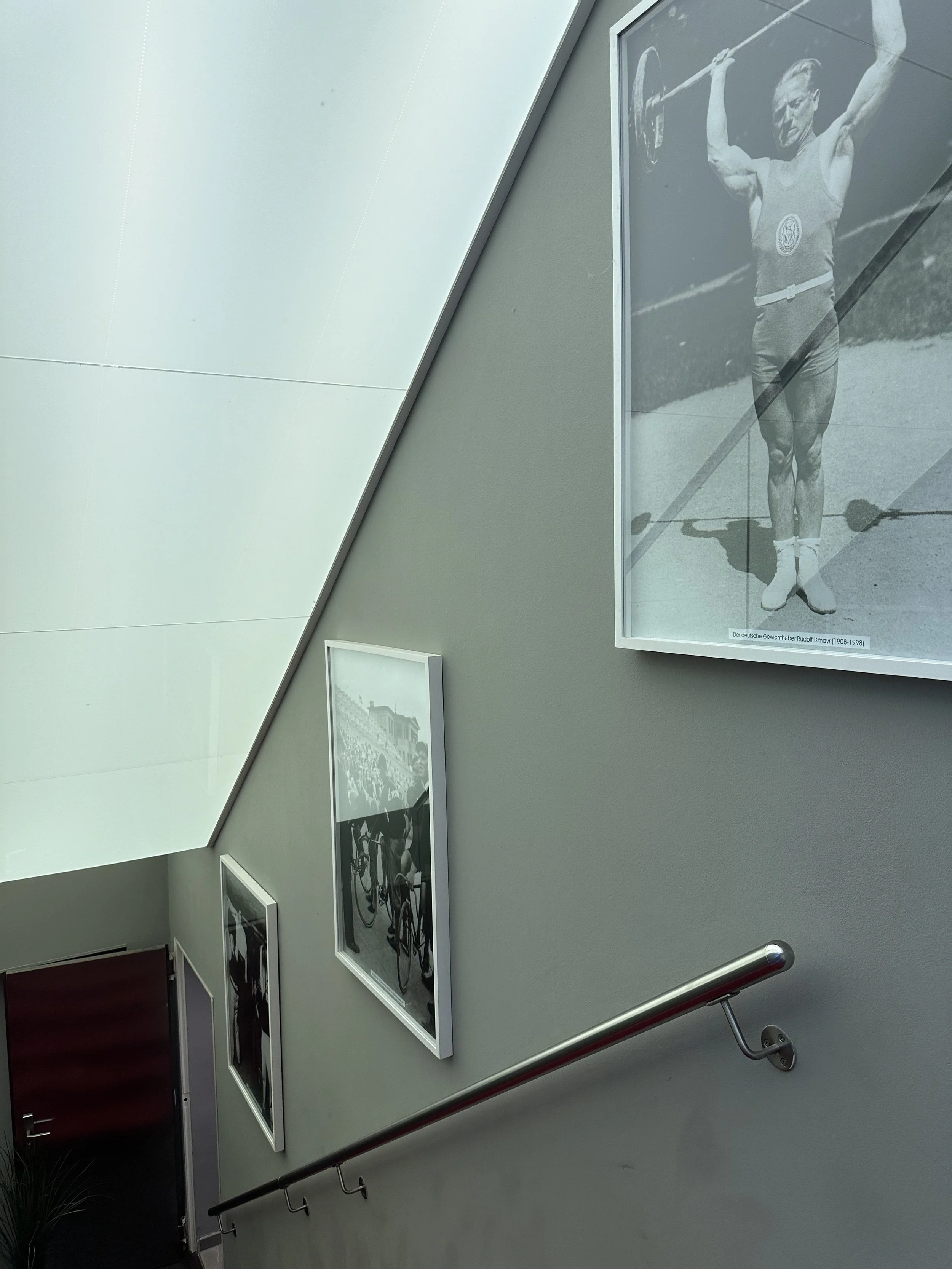 Indoor staircase with framed black-and-white photographs on the wall, including a vintage photo of a male weightlifter and a cycling race scene.