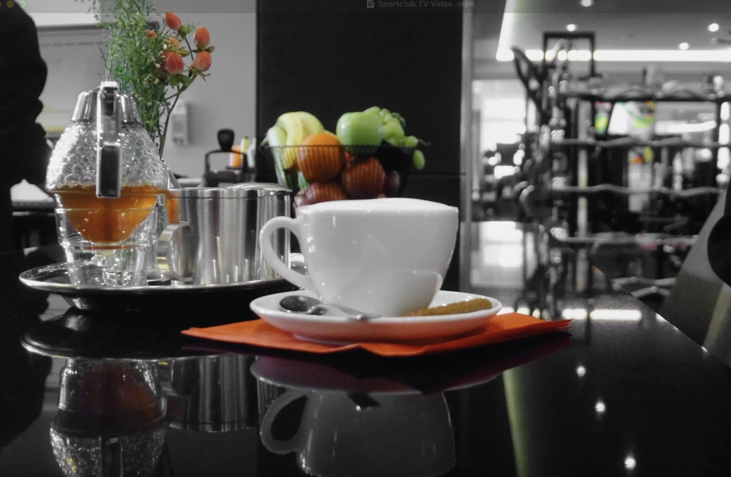 A table in a cafe with a white coffee cup on a saucer with a spoon and a sugar packet, a tray with a glass teapot, cups, and a metal pitcher, a bowl of assorted fruits in the background, and a vase with orange and pink flowers, with a blurred background showing a gym or fitness center.