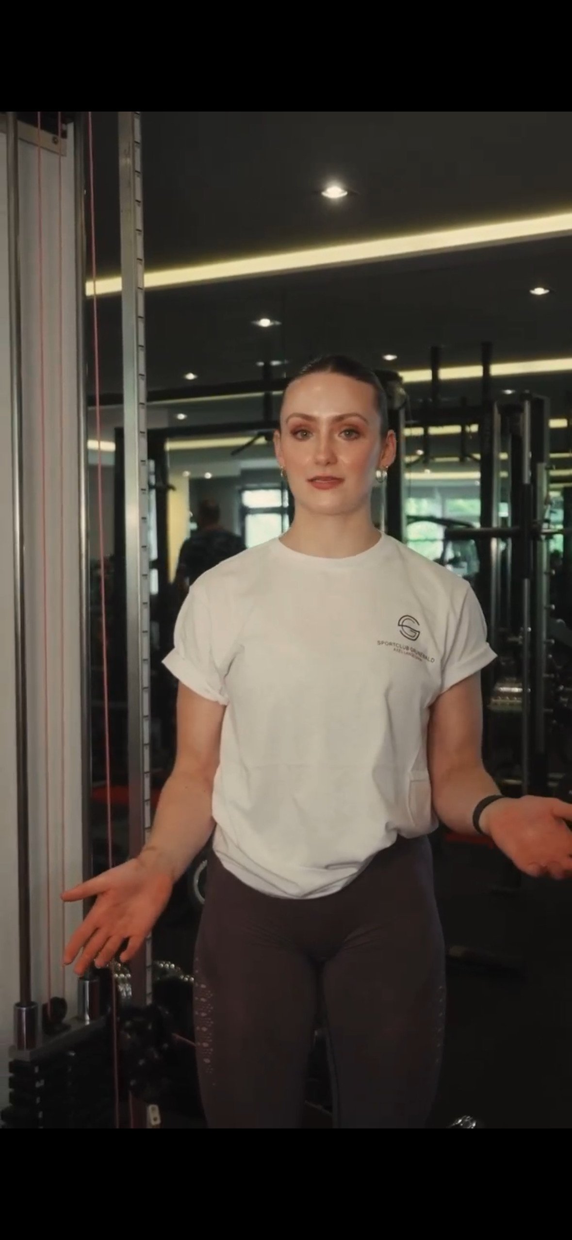 Woman standing in a gym, wearing a white T-shirt and dark leggings, with gym equipment in the background.