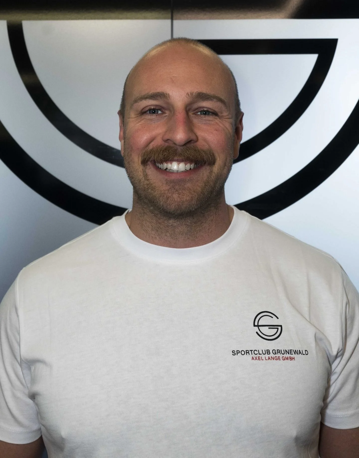 A smiling man with a beard and mustache wearing a white T-shirt with a logo and text, standing in front of a black and white abstract background.