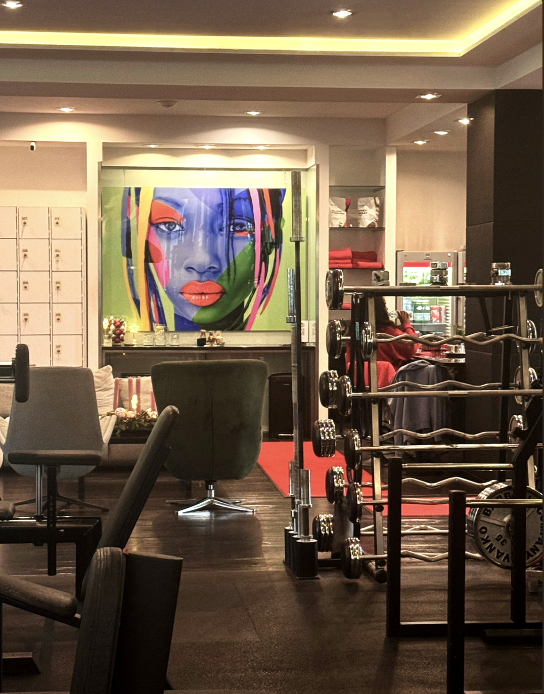 Interior of a gym or fitness center with colorful artwork of a woman's face on the wall, weightlifting equipment, and a woman working at a treadmill in the background.