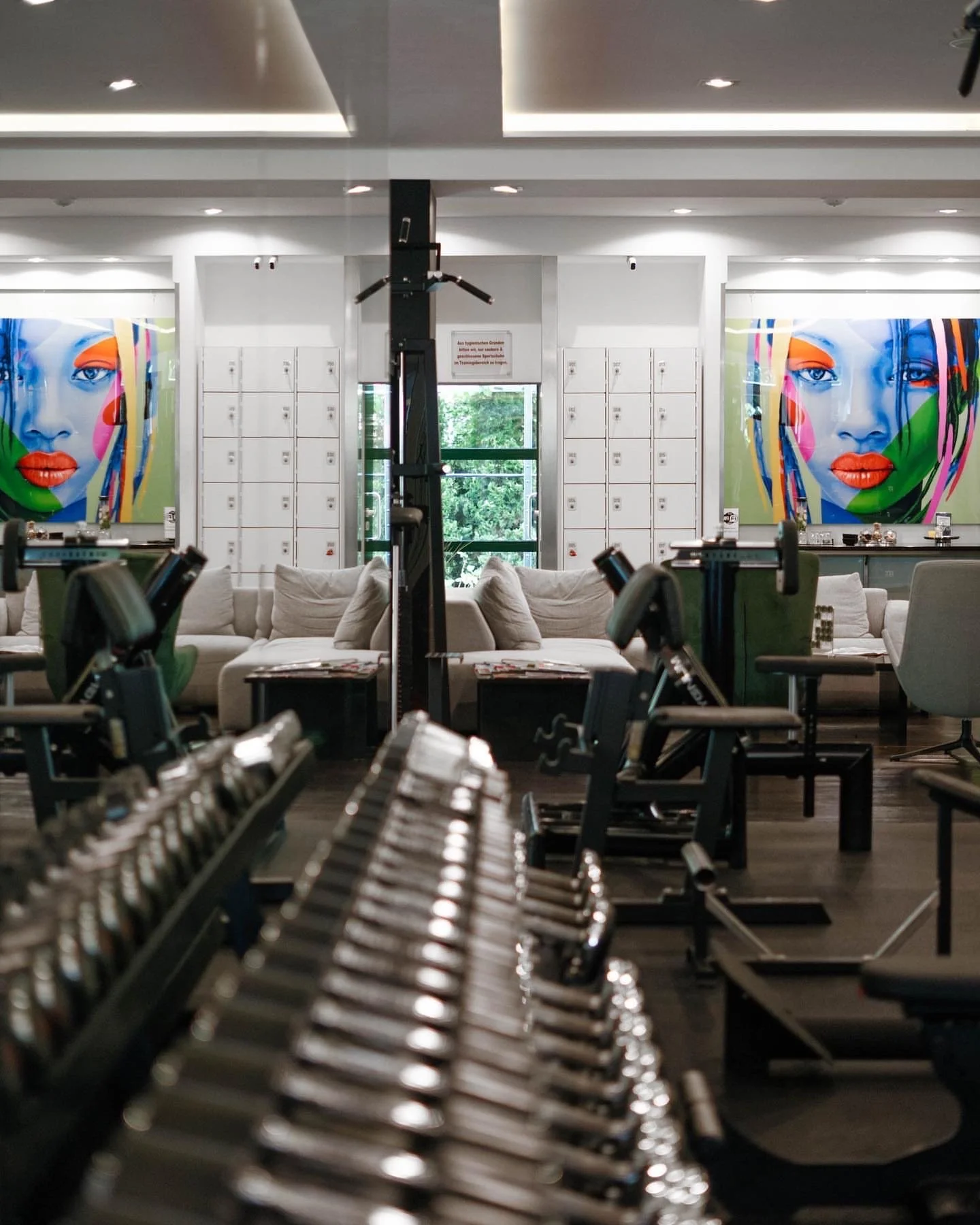 Interior of a gym with workout equipment in the foreground, white sofas and lockers in the background, and colorful artwork of a woman's face on the wall.