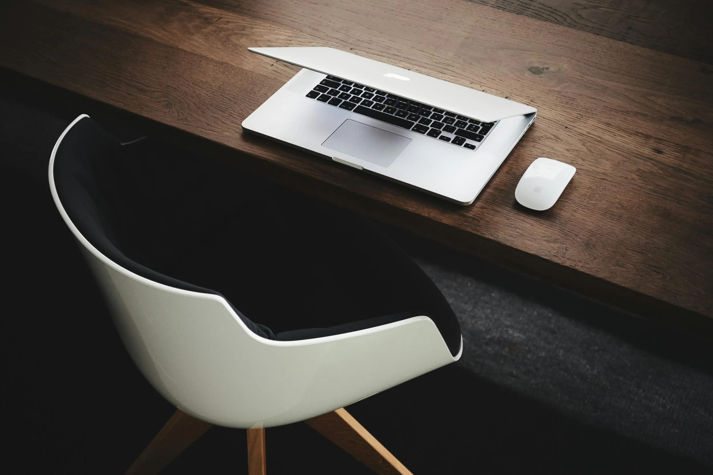 A silver MacBook laptop with a black keyboard and a large trackpad, partially open, on a wooden table. Next to the laptop is a white Apple Magic Mouse. The table is situated next to a white chair.
