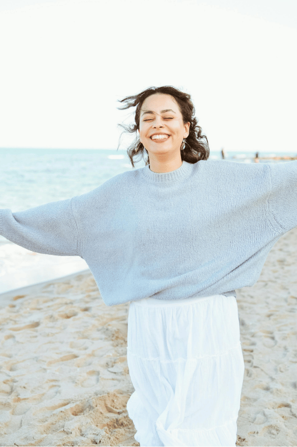 Smiling woman standing on a beach with arms outstretched, representing emotional freedom, self-acceptance, and inner peace.