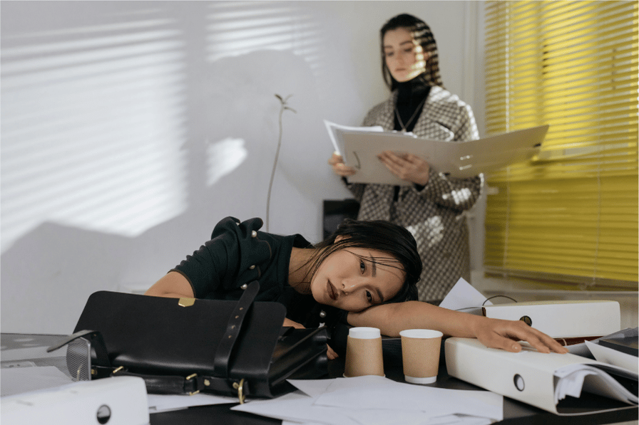 Exhausted woman resting on a cluttered desk while supervisor stands reviewing her work, representing perfectionism, overworking, and emotional exhaustion.