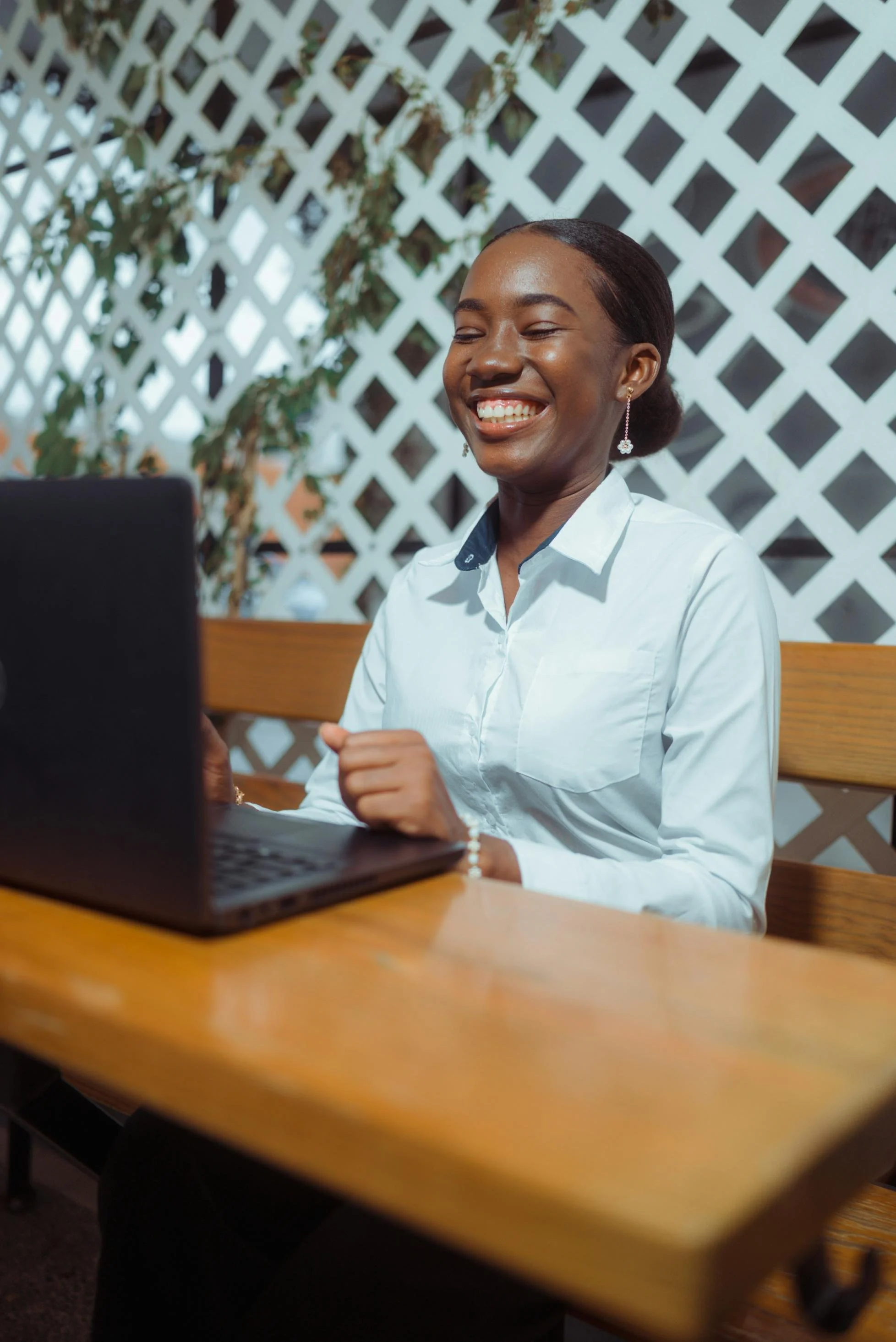 A woman with dark skin, wearing a white collared shirt and earrings, smiling while looking at a laptop. She is sitting at a wooden table with a lattice wall and plants in the background.