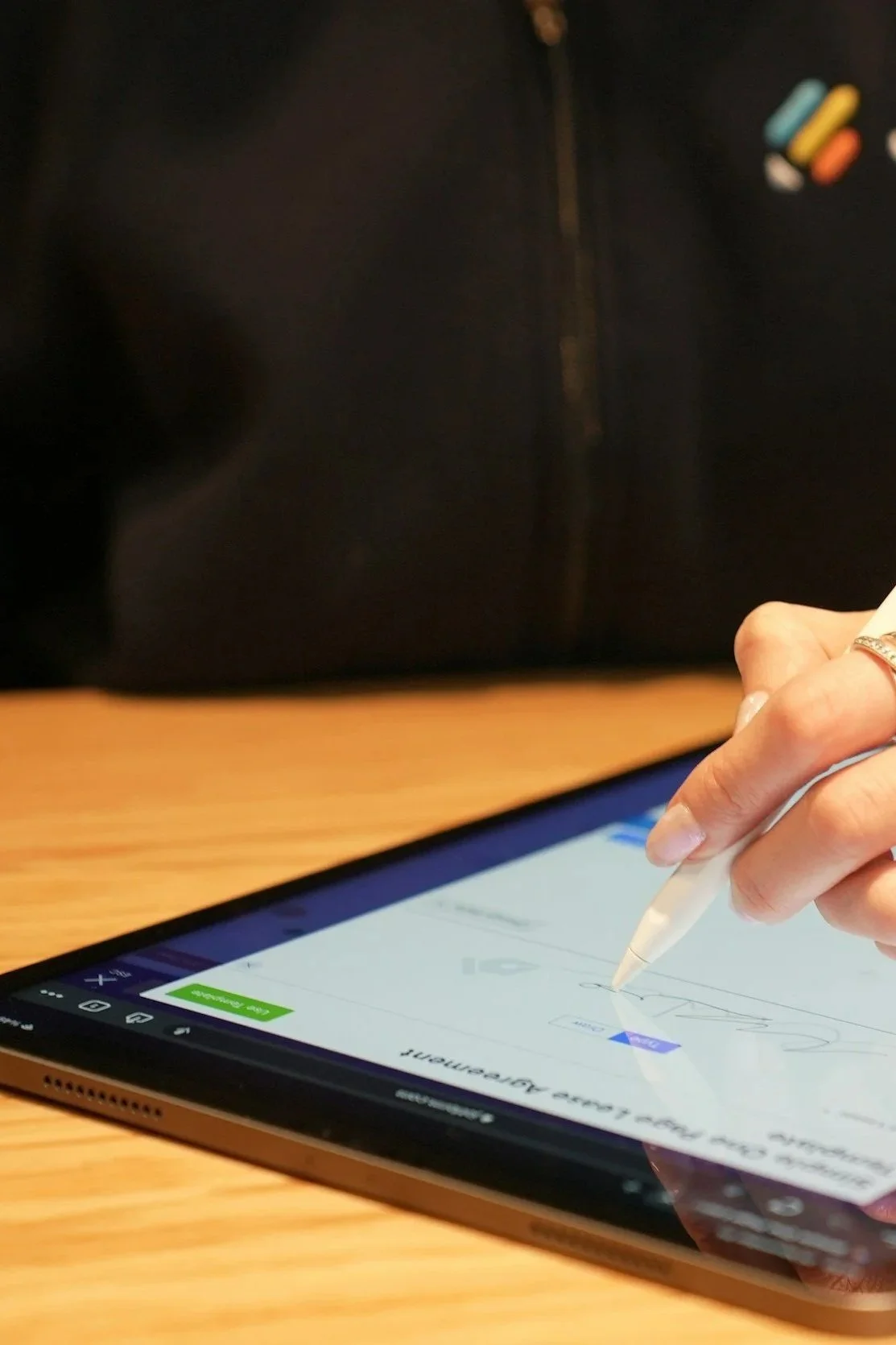 Person signing a document on a tablet device with a stylus in their right hand, on a wooden table, with a black background.