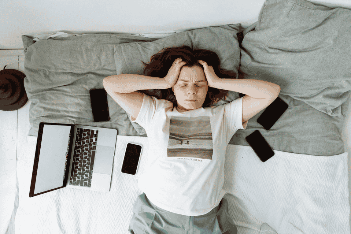 Woman lying on a bed surrounded by phones and work devices while appearing emotionally overwhelmed and mentally exhausted.
