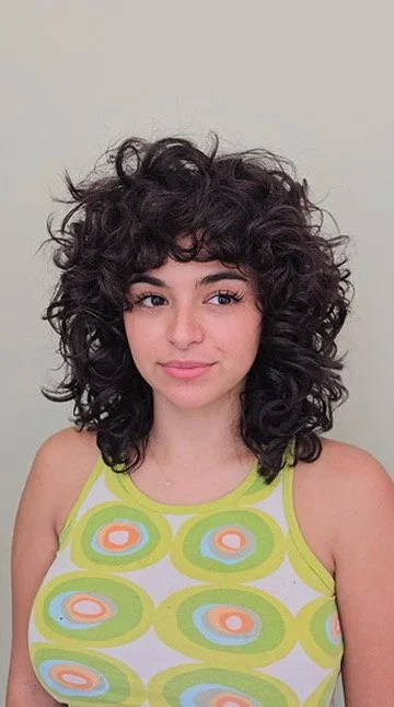 Young woman with dark, curly hair wearing a colorful sleeveless top with green, peach, and white patterns, standing against a plain light background.