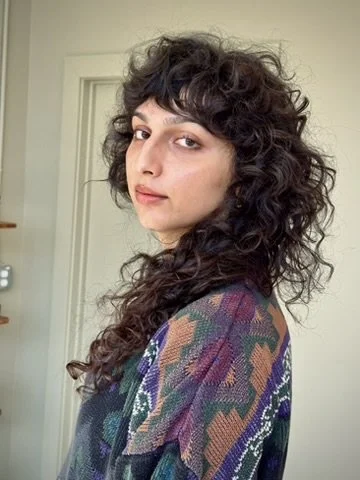 A woman with curly dark hair and fair skin looking over her shoulder in a room with a door in the background.