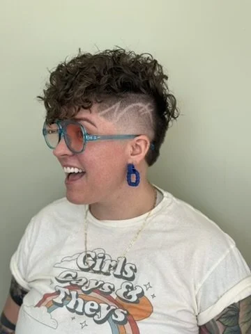 Woman with short curly hair styled in a mohawk with shaved design, wearing glasses, a white graphic t-shirt, and large blue earrings, smiling against a light green wall.