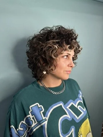 Woman with short, curly hair wearing a teal graphic T-shirt and a silver chain necklace, looking to the right against a light blue wall.