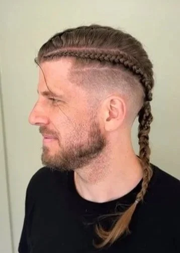 Side profile of a man with a braided hairstyle, undercut, and beard, wearing a black shirt.
