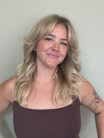 A smiling woman with blonde, wavy hair, wearing a dark tank top, standing against a light-colored wall.