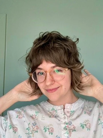 A person with short, curly brown hair wearing glasses and a floral-patterned white shirt, smiling and touching their ears.