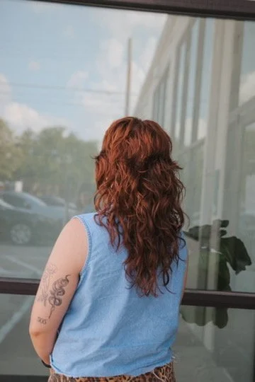A woman with curly red hair wearing a sleeveless blue top, standing indoors and looking out a window.