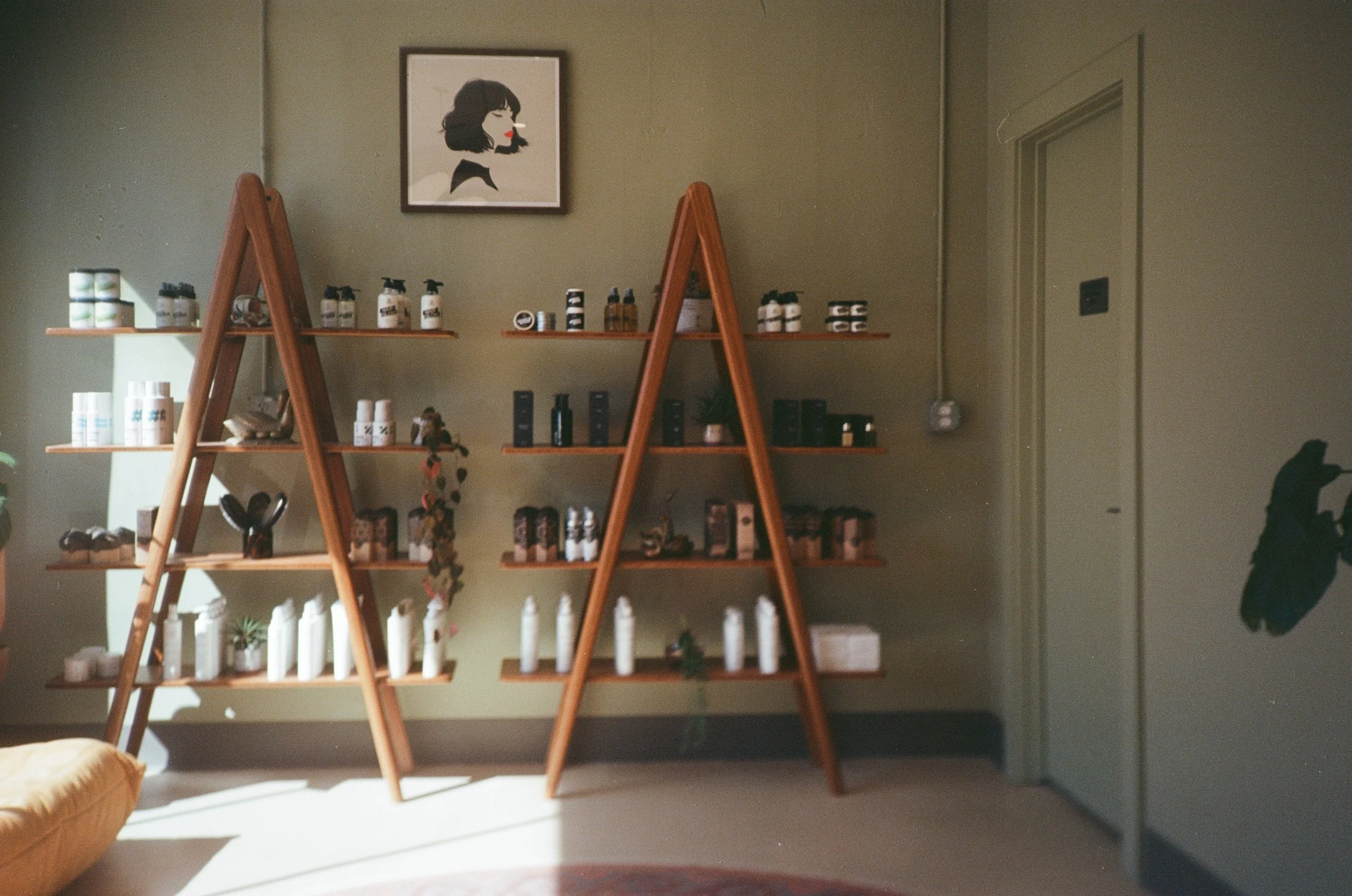A yellow sofa in a room with a light green wall, two wooden ladder shelves with products and decorative items, and a framed art print of a woman with black hair and red lips. Sunlight streams into the room.