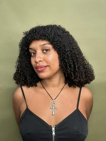 Woman with curly black hair wearing a black spaghetti strap top and an ankh necklace standing against a green background.