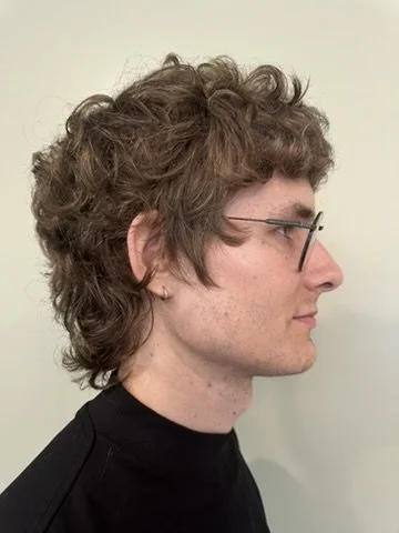 Profile side view of a young man with curly brown hair, glasses, and a black turtleneck standing against a light-colored background.