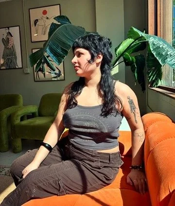 A woman with short black hair and tattoos sitting on an orange couch in a room with green plants and framed artwork on the wall.