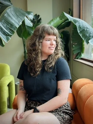 A young woman with curly hair sitting on an orange couch next to a green chair, surrounded by large banana leaves and looking out the window.