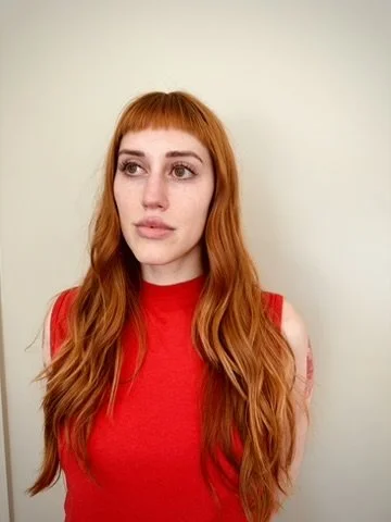 Young woman with long red hair wearing a red sleeveless top, standing against a plain light-colored background.