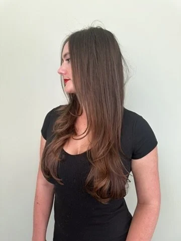 Side view of a woman with long, wavy brown hair wearing a black short-sleeve top against a plain white wall.