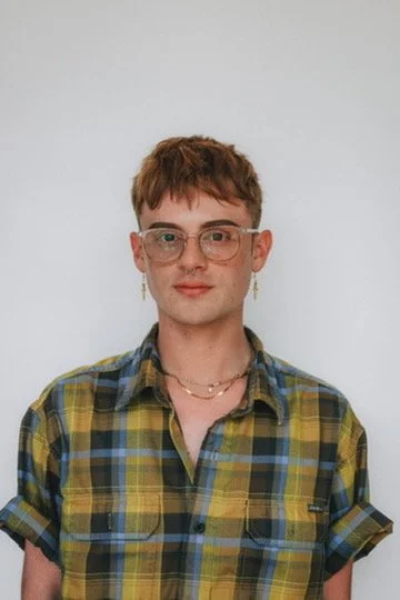 Young person with short brown hair, glasses, and earrings, wearing a yellow, green, and blue plaid shirt with rolled-up sleeves, standing against a plain white background.