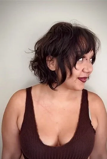 A woman with short, wavy, dark brown hair with bangs, wearing a dark brown sleeveless top, looking to her right with dark lipstick and a slight smile, standing against a plain light-colored background.