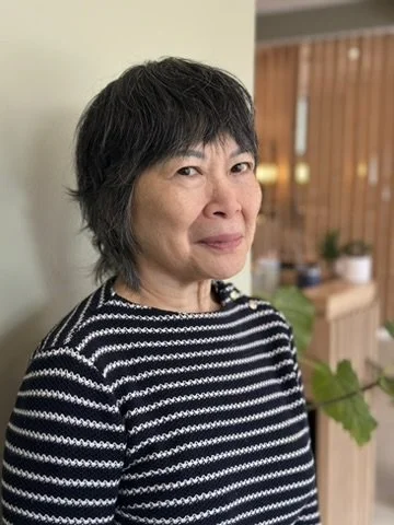 A woman with short black hair wearing a black and white striped shirt, standing indoors with a wooden partition and some plants in the background.
