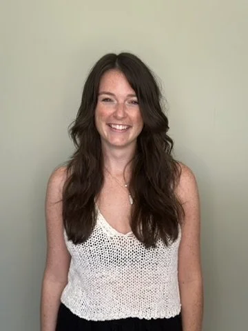 A young woman with long, wavy dark hair, smiling, wearing a white sleeveless knit top, standing against a light green background.