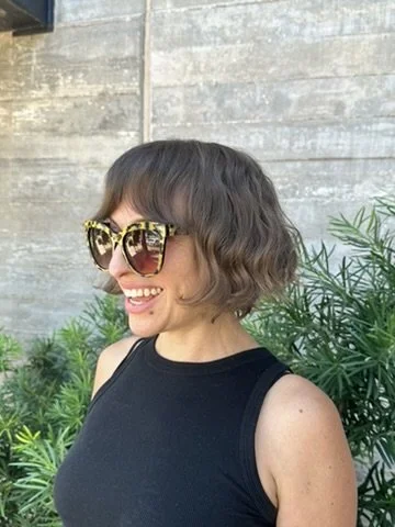 A woman with short, wavy hair wearing big, patterned sunglasses smiling outdoors, with green plants and a concrete wall in the background.