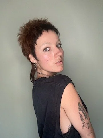 Person with short, spiked hair and tattoos on their arm, wearing a black sleeveless top, standing against a plain light green background.
