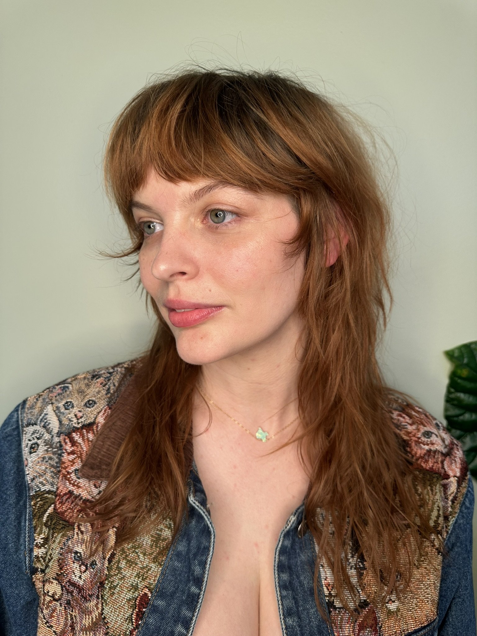Close-up of a woman with red wavy hair and green eyes, wearing a colorful tapestry jacket and a delicate necklace with a pendant, standing against a light-colored wall.