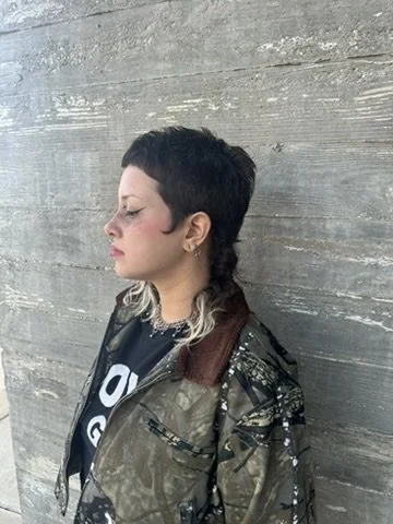 Profile of a person with short black hair and blond tips, wearing a camouflage jacket and a black graphic T-shirt, standing against a wooden wall.