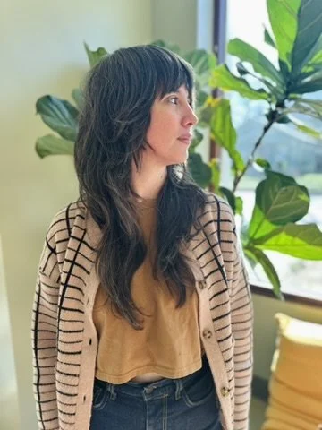 Profile of a woman with long dark wavy hair, wearing a beige striped cardigan over a yellow top, standing indoors near large green plants.