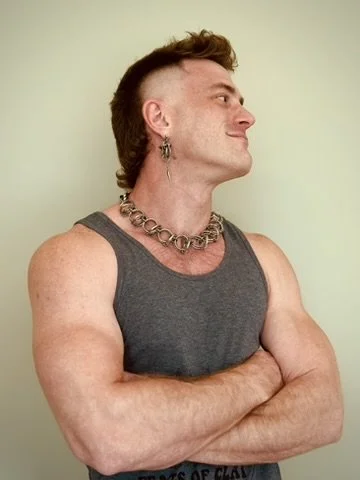 Side profile of a muscular person with styled hair, wearing a sleeveless gray top, stud earring, and a chain necklace, standing with arms crossed against a plain light-colored background.