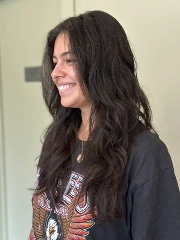 Side profile of a smiling woman with long, wavy dark hair wearing a black t-shirt with a graphic design, standing indoors.