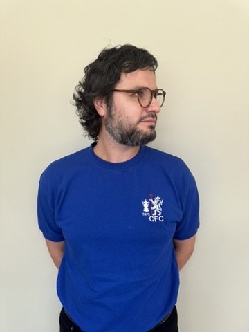 A man with curly dark hair and glasses wearing a blue T-shirt with a small logo on the chest standing against a plain light-colored wall.