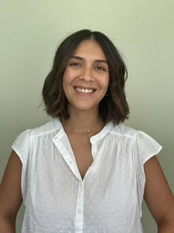 A woman with shoulder-length dark hair smiling, wearing a white blouse, standing against a light green background.