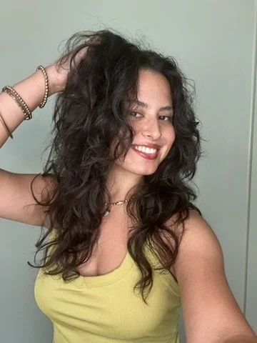 A woman with curly dark hair wearing a yellow top and jewelry, smiling and taking a selfie.
