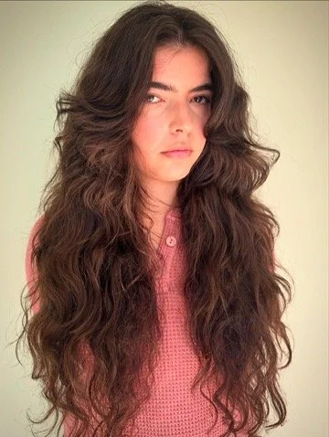 Young woman with long, wavy brown hair wearing a pink top, looking at the camera.