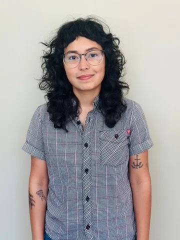 Young woman with curly dark hair, glasses, wearing a gray checkered short-sleeve shirt, standing against a plain wall.